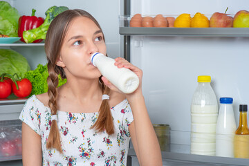 Beautiful young teen girl holding bottle of milk and drinks while standing near open fridge in kitchen at home