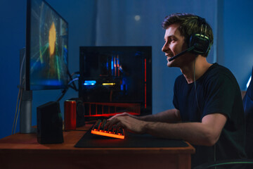 Young handsome pro gamer playing in online video game on computer at home using headphones and microphone for talking with his team, smiling, enjoying the play. Side view. Cybersport concept © Arsenii