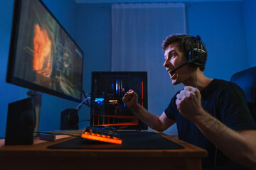 Cybersport young pro gamer happy with winning the game, feel exited, show YES hand gesture, celebrates victory in online game competition. Side view. Guy playing video game at home in his room - obrazy, fototapety, plakaty