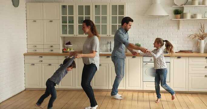 Young couple and little cute kids dancing. Parents and children hold hands jumping barefoot on warm wooden floor in kitchen, enjoy active carefree weekend celebrate move day to new modern home concept