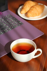 White cup with black tea and croissants in a saucer on the work table. Remote work at home. Snack near the laptop. Sweet morning.