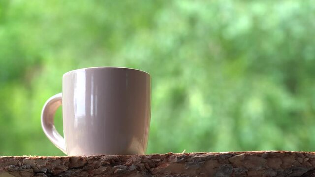 Closeup view 4k stock video footage of brown cup of tea or coffee drink standing on brown organic wooden desk with green morning summer or spring bokeh background seen behind mug with copyspace