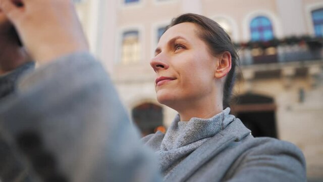 Woman in a coat standing in the middle of an old square, using smartphone and taking a photo.