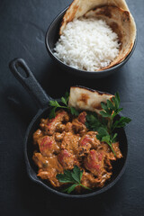 Cast-iron skillet with indian butter chicken masala and a bowl of white rice, vertical shot on a black stone surface