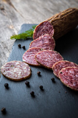 Sliced salami sausage on a board. Meat product and herbs on a dark back. A quick meal or snack. Dried sausage with black peppercorns.