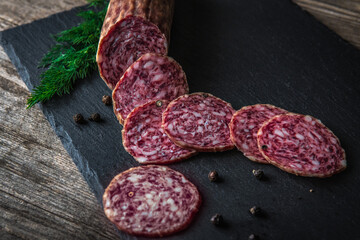 Sliced salami sausage on a board. Meat product and herbs on a dark back. A quick meal or snack. Dried sausage with black peppercorns.
