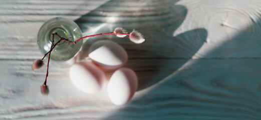 A sprig of willow with fluffy silver buds in a glass bottle and white eggs on the white table at...