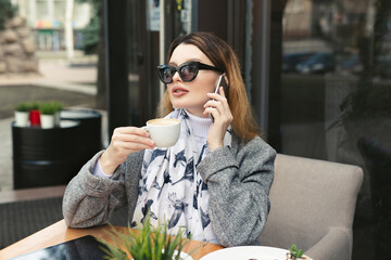 Beautiful young woman in black sunglasses is drinking coffee in a cafe on the street and talking on a mobile phone