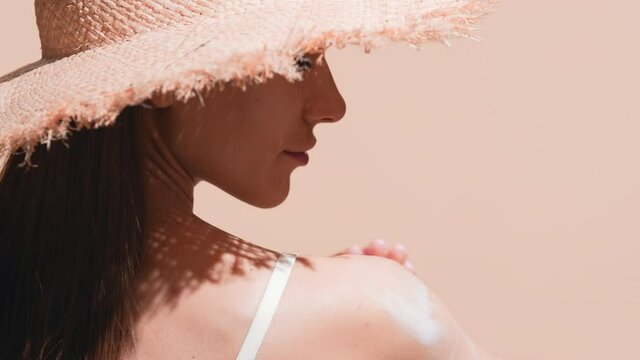 Young gorgeous European woman with long dark hair in a straw hat thoroughly applies sunscreen on her shoulder against beige background | Sunscreen applying shot for body care commercial