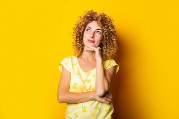 curly young woman holds her hand under her chin looks thoughtfully upwards on a yellow background