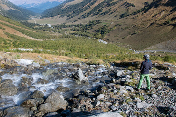 A tourist (man with trekking poles) stands on the bank of mountain stream and looks at Sophia river valley on sunny autumn day. Arkhyz, Caucasus, Russia.