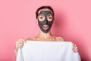 surprised young woman covers herself with a towel with a clay mask on her face on a pink background