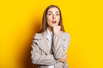 Surprised Young woman in a coat holds a hand under her chin on a yellow background. Banner