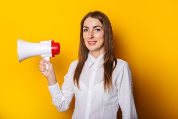 Young woman in a white shirt holds a megaphone in her hands on a yellow background. Hiring concept,...