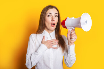 Young woman with a surprised face in a white shirt holds a megaphone in her hands on a yellow background. Hiring concept, help wanted. Banner