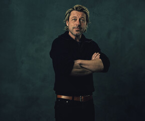 Relaxed blonde man with stubble beard in black wool sweater and jeans in front of a dark wall.