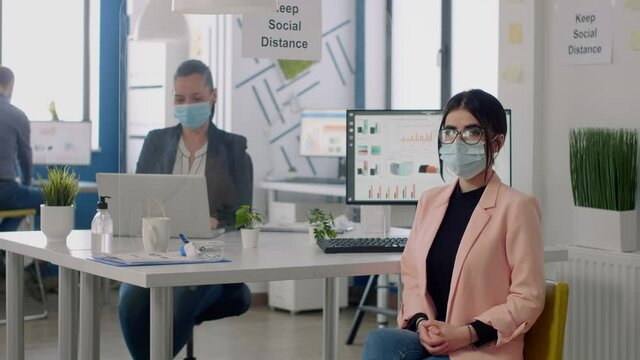 Portrait of manager with face mask sitting on chair at desk table in nre normal business company office. Teamworkers working in background respecting social distancing during coronavirus pandemic