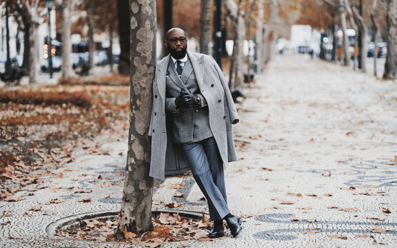 A Dapper Black Senior Entrepreneur In Eyeglasses And An Elegant Outfit With A Custom-made Costume And A Coat, Is Leaning Against A Tree On An Autumn Boulevard Alleyway; A Copy Space Place On The Right