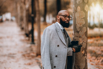 A street portrait with a shallow depth of field and selective focus on a dapper bearded black man entrepreneur in an elegant coat, gloves, eyeglasses, and a suit, near the tree of a fall alleyway