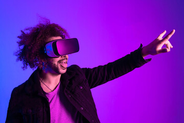 Young model with beard and afro hair in virtual reality glasses on lilac and blue background making victory sign. Augmented reality, future technology concept. VR. Neon light.