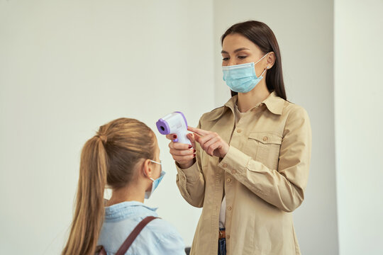 Attractive young female teacher wearing protective mask screening schoolgirl using digital thermometer while kid coming to school