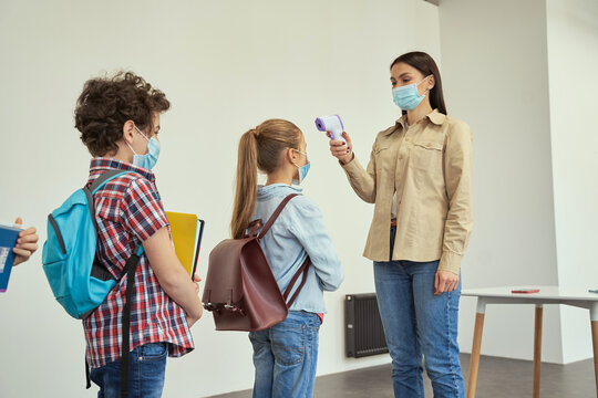 Female teacher wearing protective mask screening school children for fever against the spread of Covid19, using digital thermometer while kids coming to school - Powered by Adobe