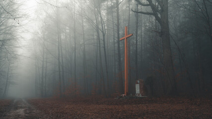 Pomnik z krzyżem przy drodze w mglistym lesie.
