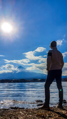 Fototapeta premium A man standing at the side of Kawaguchiko Lake, Japan with the view on Mt Fuji. The magnificent mountain surrounded by clouds. Soft reflections in the calm surface of the lake. Serenity and calmness