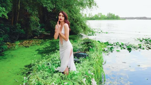 Fantasy happy brunette woman fashion model posing in boat, sexy white dress, slits on long legs. Fairy Girl princess. summer nature blue water lake green tree. Spring fresh plants. beauty river nymph