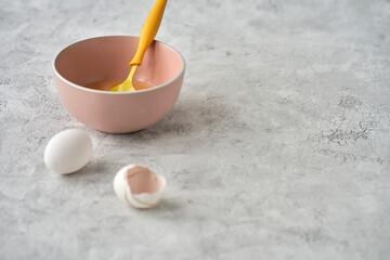 A bowl with egg mass on a gray textured background. Next to it is a chicken egg and an egg shell. Selective focus. Copy space.