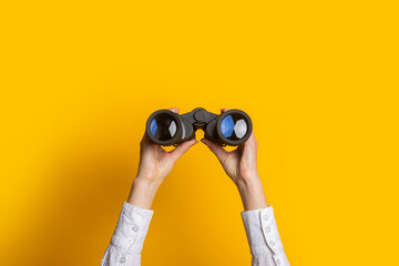 female hands hold black binoculars on a bright yellow background - obrazy, fototapety, plakaty