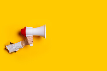 Hand holds a megaphone from a hole in the wall on a yellow background. Concept of hiring, advertising something. Banner - obrazy, fototapety, plakaty