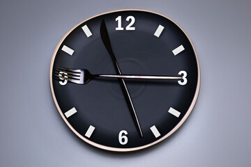 Clock on the black plate, with cutlery, grey background. The concept of intermittent fasting, lunchtime, diet and weight loss.