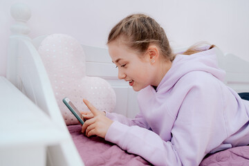 A Caucasian teenage girl uses a smartphone while resting on a bed in her room.