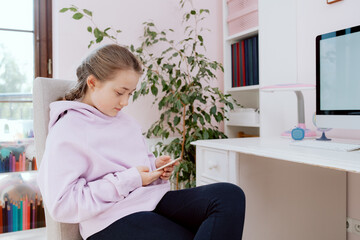A Caucasian teenage girl uses a smartphone while sitting on a chair at a desk in her room. The girl browses content on the Internet. She posts on social media.