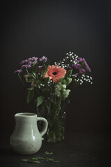 Vertical still life with a mix of spring flowers. An orange gerbera, red carnation, purple chrysanthemums and white gypsophila in a glass vase. Porcelain milk jug. Dark moody studio shot, copy space