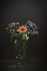 Vertical still life with a mix of spring flowers. An orange gerbera, red carnation, purple chrysanthemums and white gypsophila in a glass vase. Dark moody studio shot with copy space.
