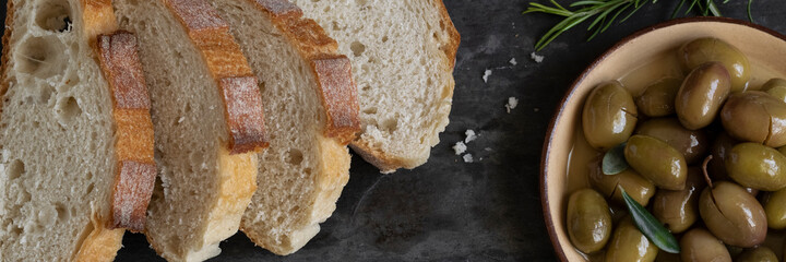 Italian sliced ciabatta bread on chopping board with herbs, extra virgin oil and marinated olives on dark grunge backdrop