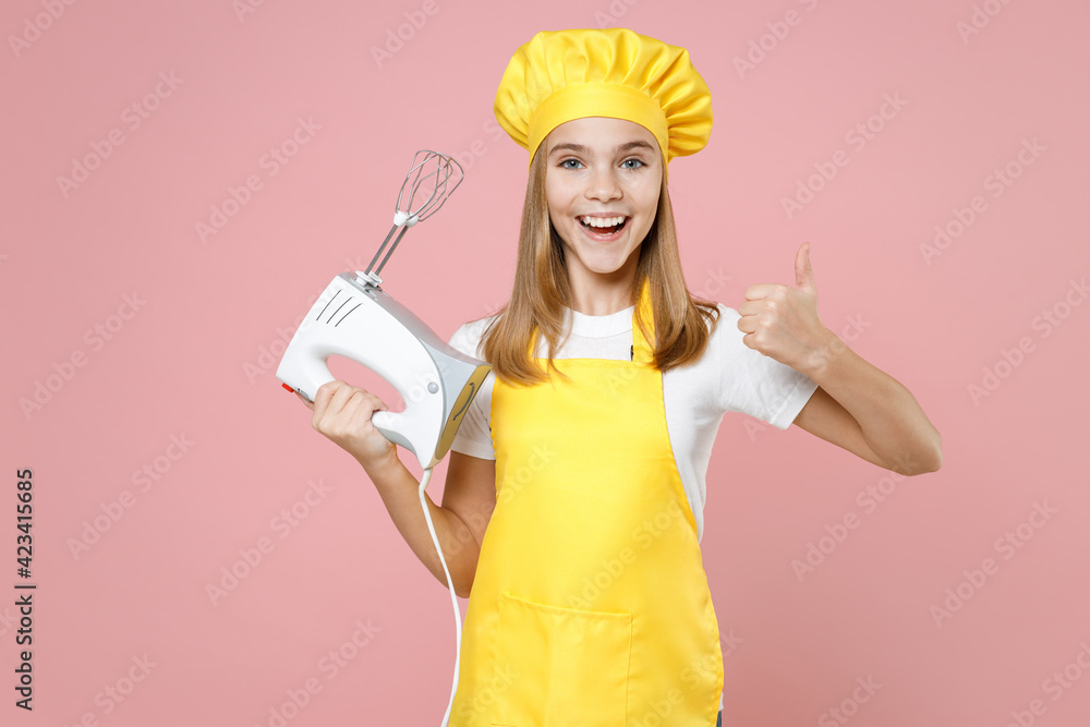 Wall mural Teen smiling girl chef cook confectioner housewife mother's helper baker in yellow apron white t-shirt cap hold kitchen mixer show thumb up like gesture isolated on pink background Food cake concept.