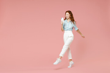 Fototapeta na wymiar Full length young student smiling redhead woman 20s wearing blue shirt pants using mobile cell phone chatting online in social network walking go isolated on pastel pink background studio portrait.