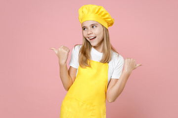 Teen girl chef cook confectioner baker housewife mother's helper wear yellow apron white t-shirt cap point thumb finger aside on workspace area isolated on pink background studio. Food cake concept.