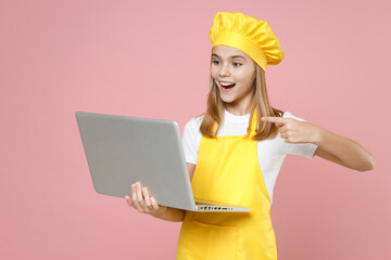 Teen surprised girl chef cook confectioner housewife baker wearing yellow apron white tshirt cap point index finger on laptop pc computer isolated on pink background studio portrait Food cake concept