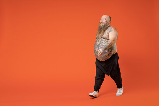 Full Length Side Profile View Of Fat Pudge Obese Chubby Overweight Tattooed Bearded Man 30s Has Big Belly In Black Pants Naked Torso Walking Going Stroll Isolated On Orange Background Studio Portrait.