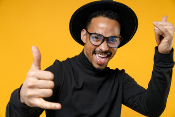 Young smiling happy fun african american man 20s wearing stylish black hat shirt eyeglasses show close up shaka greeting gesture stretch hands to camera isolated on yellow background studio portrait