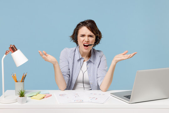 Young indignant operator employee business woman in shirt set microphone headset helpline assistance sit work at office desk with pc laptop spreading hands isolated on pastel blue background studio.