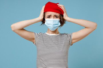 Young troubled sad woman 20s wearing french beret red hat striped t-shirt sterile face mask from coronavirus covid-19 pandemic quarantine hold head isolated on pastel blue background studio portrait.