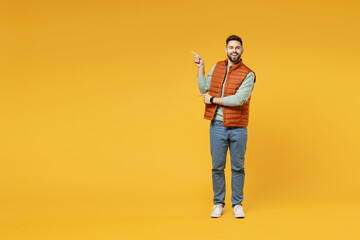 Full length young smiling happy confident smiling cheerful fun caucasian man in orange vest mint sweatshirt point index finger aside on copy space area mock up isolated on yellow background studio