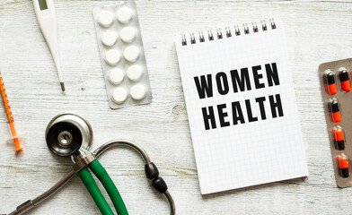 WOMEN HEALTH is written in a notebook on a white table next to pills and a stethoscope.