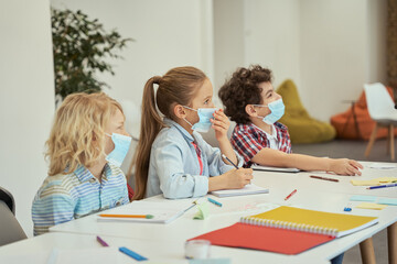 Fototapeta premium Group of elementary school kids wearing protective mask during coronavirus pandemic listening to teacher while sitting together at the table in a classroom