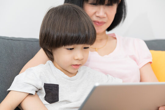 Cheerful Asian mother with little son sitting on sofa and playing video game on tablet while spending time together at home.